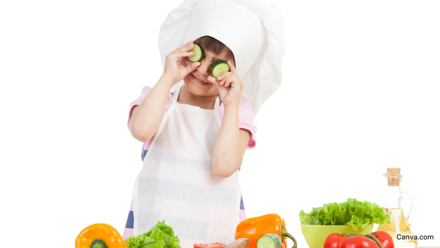 enfant tenant deux rondelles de concombre devant ses yeux, il porte un tablier et un bonnet de chef et il y a des légumes colorés devant lui